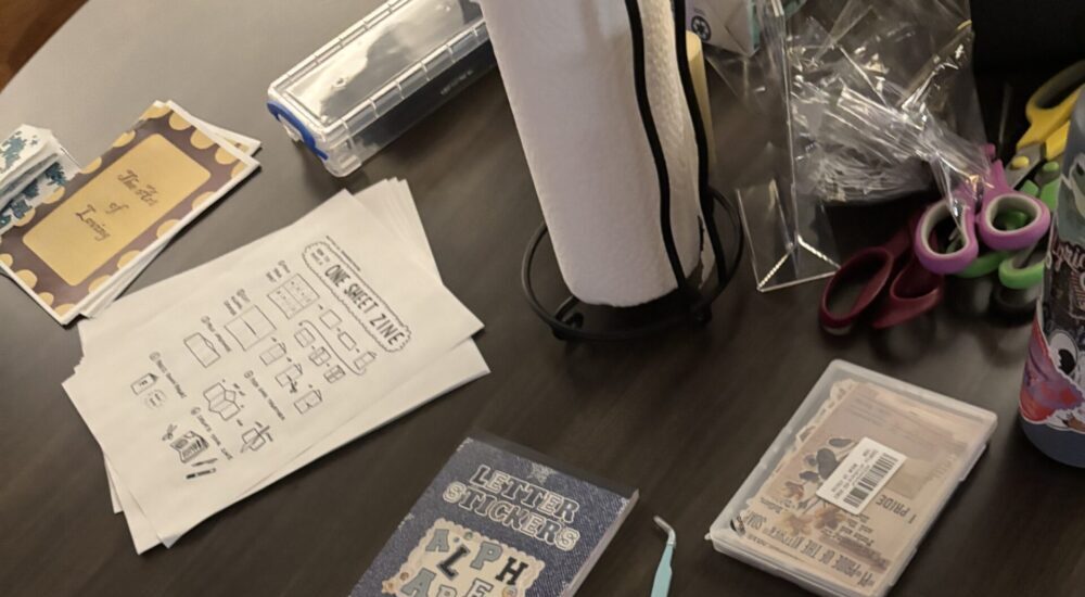 Overhead view of a dark wooden table cluttered with paper cutouts, stickers, small notebooks, and craft supplies.