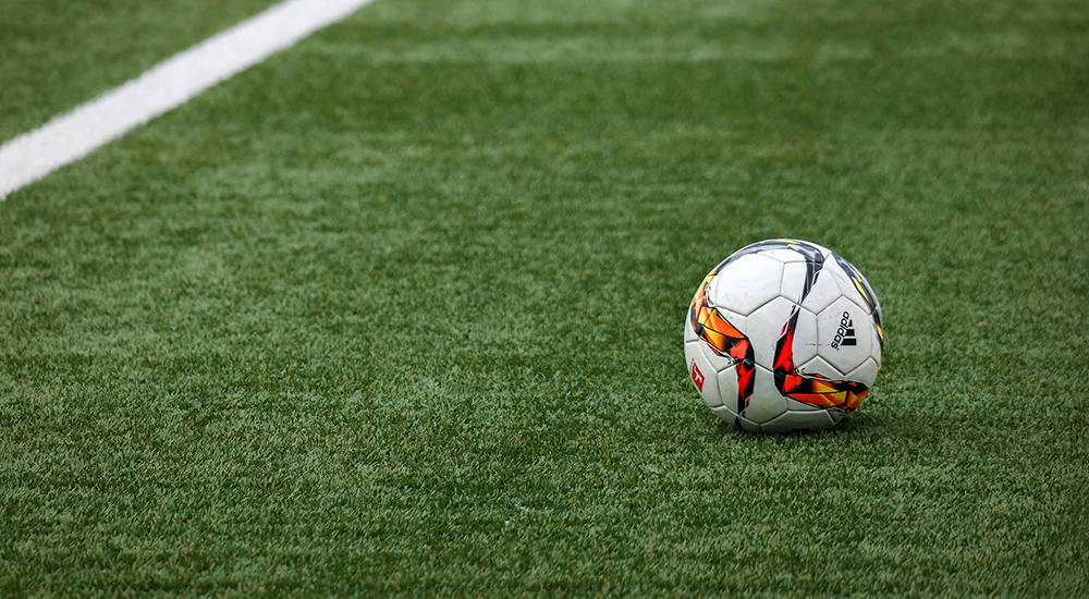 A close-up shot of a white Adidas soccer ball with orange and black geometric graphics resting on a green turf field near a thick white boundary line.