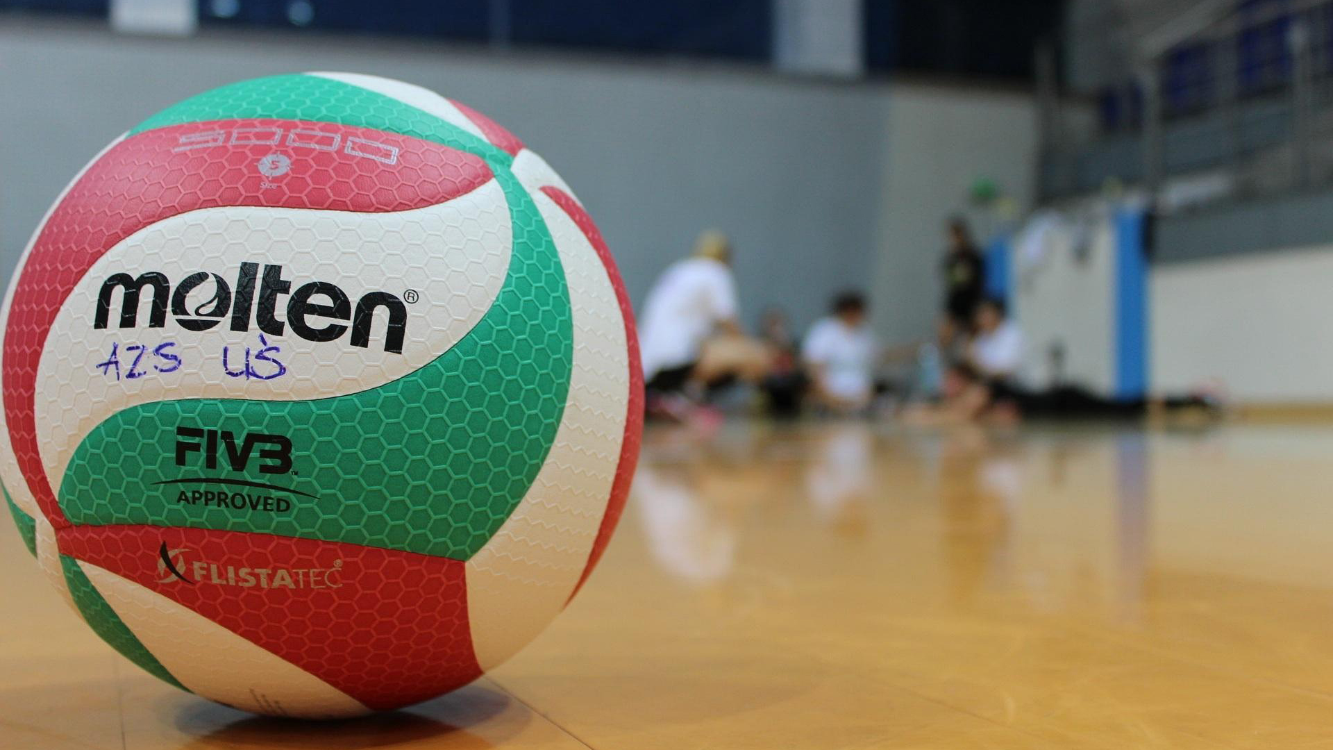 A close-up shot of a red, green, and white Molten volleyball resting on a wooden court floor, with blurred figures sitting in the background.