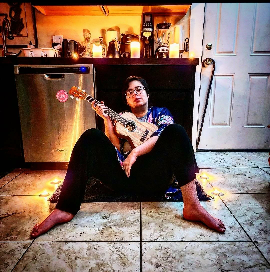 A person sitting cross-legged on a tiled kitchen floor while playing a ukulele.