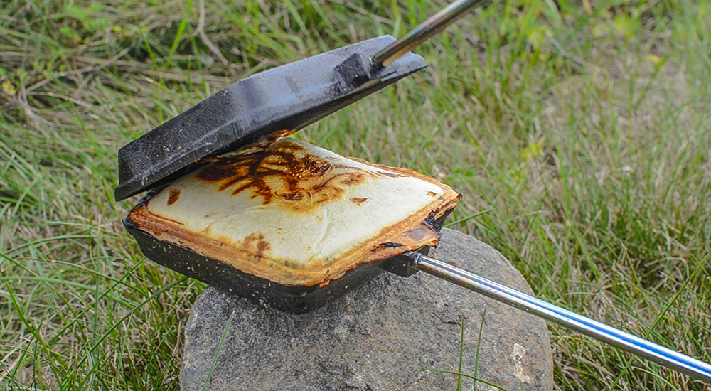 A open cast-iron pie iron resting on a rock in a grassy outdoors area, revealing a golden-brown toasted pudgie pie sandwich with charred edges inside.