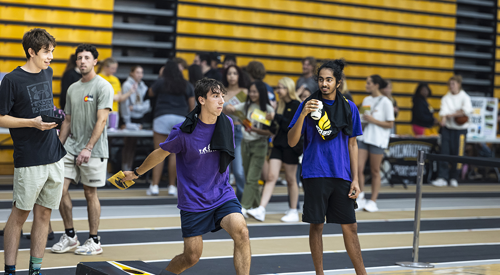 A young man in a purple t-shirt tossing a yellow beanbag while playing cornhole indoors on a track with yellow and black bleachers and other participants in the background.