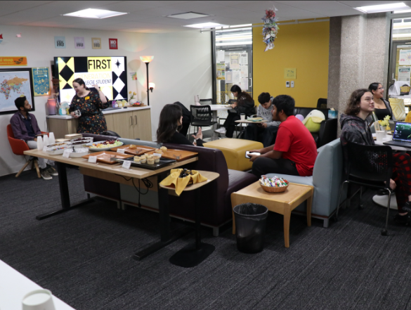 A indoor gathering inside a student resource center with carpeted floors and modern lounge furniture. Several students and staff members are sitting on couches, chairs, and at tables, chatting and using laptops. A long table in the center is set up with plates of food, pastries, and drinks, next to a wall display that reads "FIRST+."
