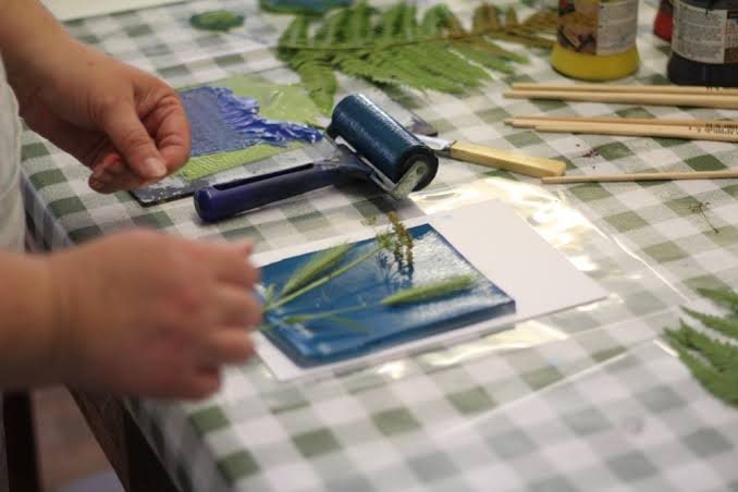 A close-up view of a person's hands placing pressed leaves and botanical cuttings onto a gel printing plate prepped with dark blue ink. Beside the plate sits a blue brayer, wooden craft sticks, extra foliage, and paint jars on a green and white checkered tablecloth.