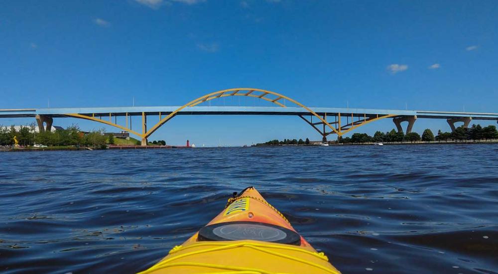 A view from the bow of a yellow kayak paddling across open water toward a large tied-arch bridge under a clear blue sky.