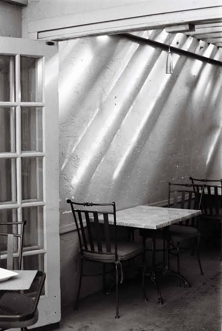 A black and white photograph showing a small patio dining setup next to an open glass paneled door. Diagonal streaks of sunlight cast shadow patterns across a wall behind a small square table paired with wrought-iron chairs.