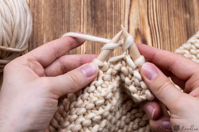 Close-up view of two hands holding wooden knitting needles, working a knit stitch into a piece of thick, cream-colored wool fabric against a wooden surface.