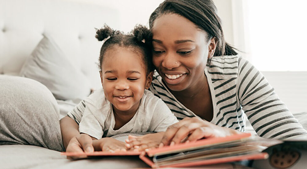A smiling Black woman and a young child lie side-by-side on a bed, looking down together at an open picture book.