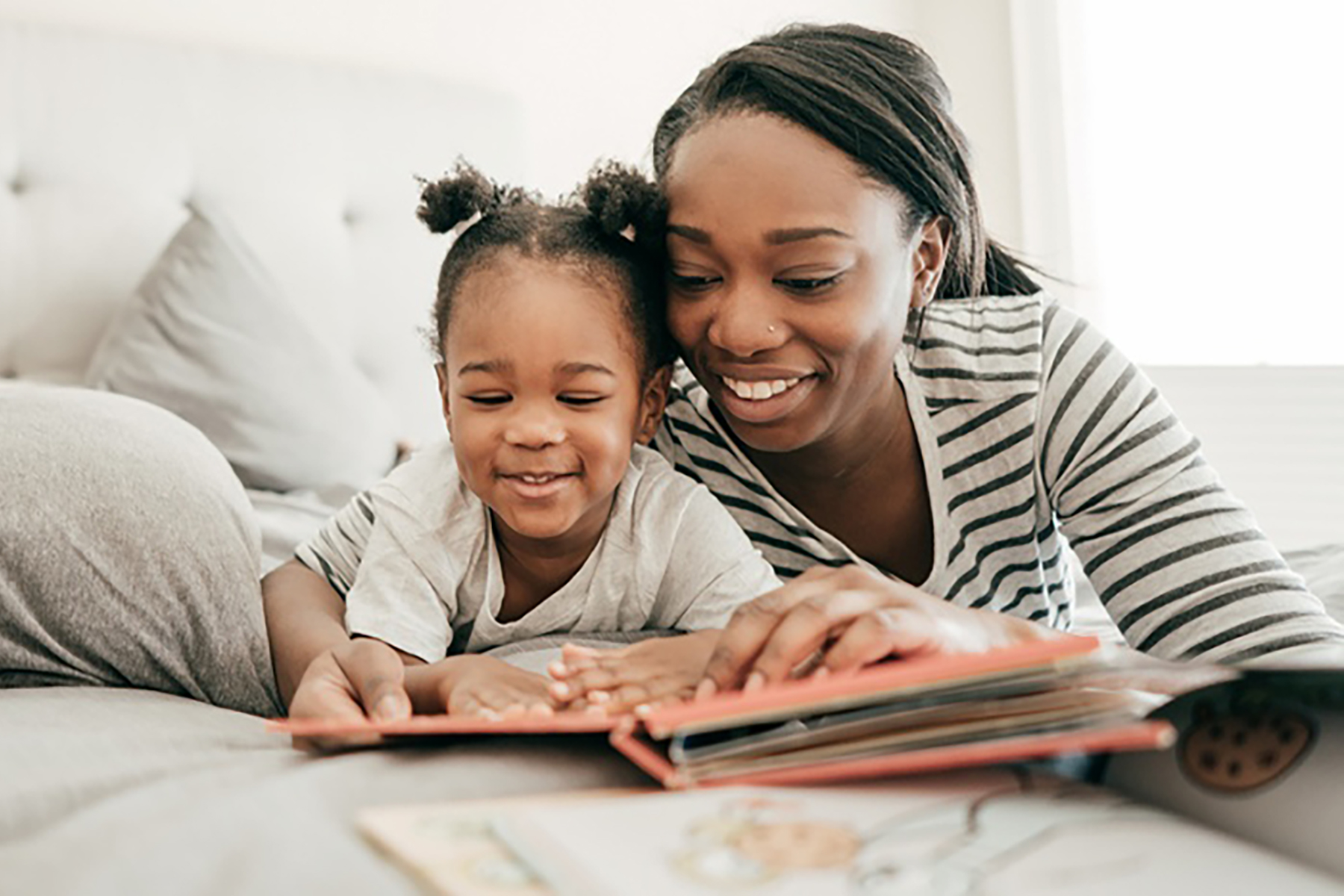 A smiling Black woman and a young child lie side-by-side on a bed, looking down together at an open picture book.