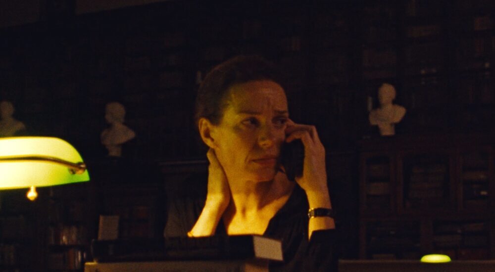 A person sits at a dimly lit desk, speaking on a landline phone while lit by the warm yellow glow of a banker's lamp amidst stacks of books.