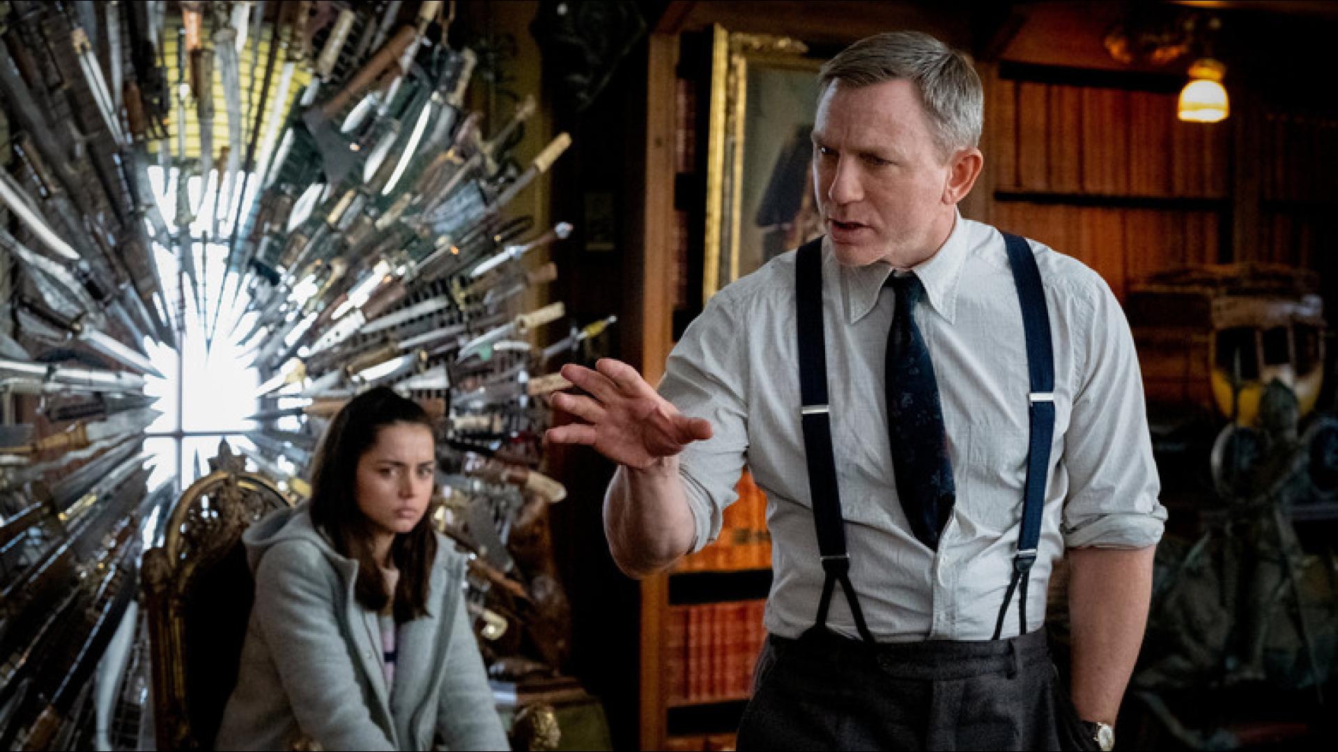 Daniel Craig as detective Benoit Blanc gesturing while speaking to Ana de Armas's character Marta in front of a circular display of knives from the movie Knives Out.