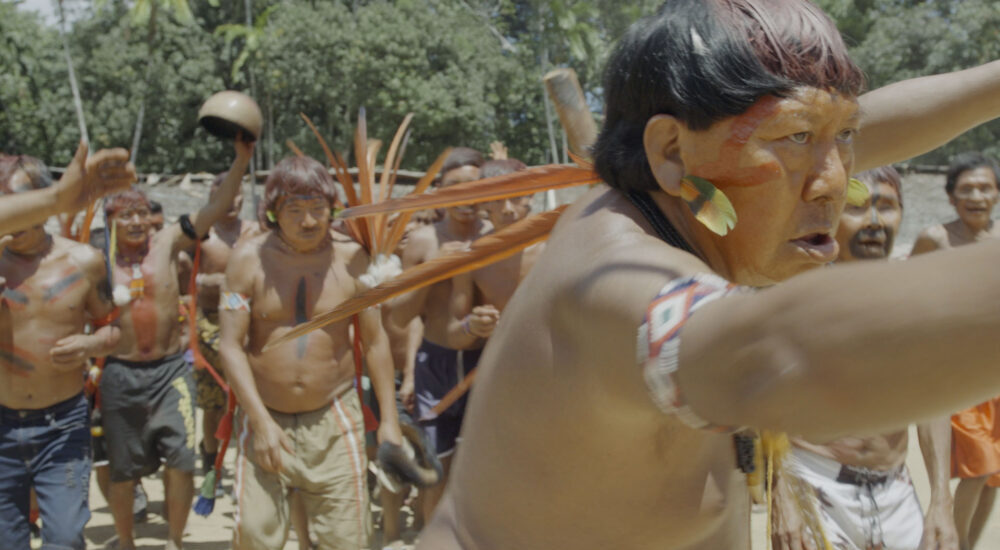 An Indigenous man with red facial paint and arm bands stands with arms outstretched in the foreground, leading a group of Indigenous people gathering outdoors amidst trees.