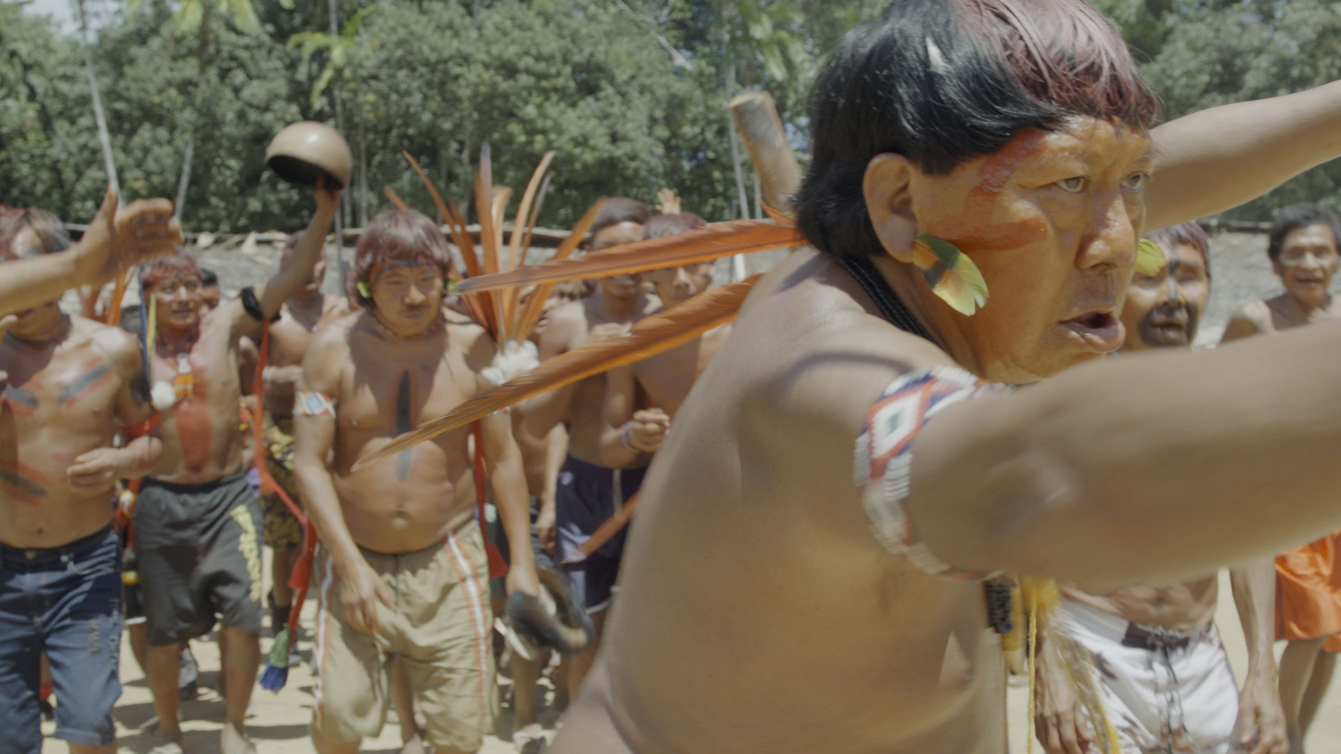An Indigenous man with red facial paint and arm bands stands with arms outstretched in the foreground, leading a group of Indigenous people gathering outdoors amidst trees.