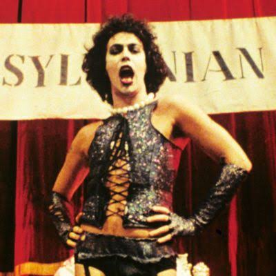 Tim Curry as Dr. Frank-N-Furter in The Rocky Horror Picture Show, wearing a glittery corset and elbow-length gloves with hands on hips, standing in front of red curtains and a banner reading "TRANSYLVANIA".