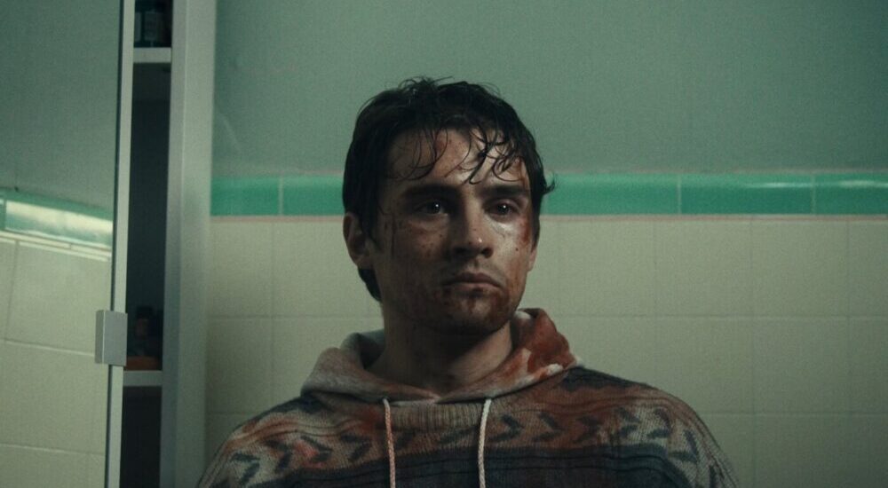A man with dark, wet hair and dirt on his face stands in front of a tiled wall wearing a patterned zip-up hoodie.