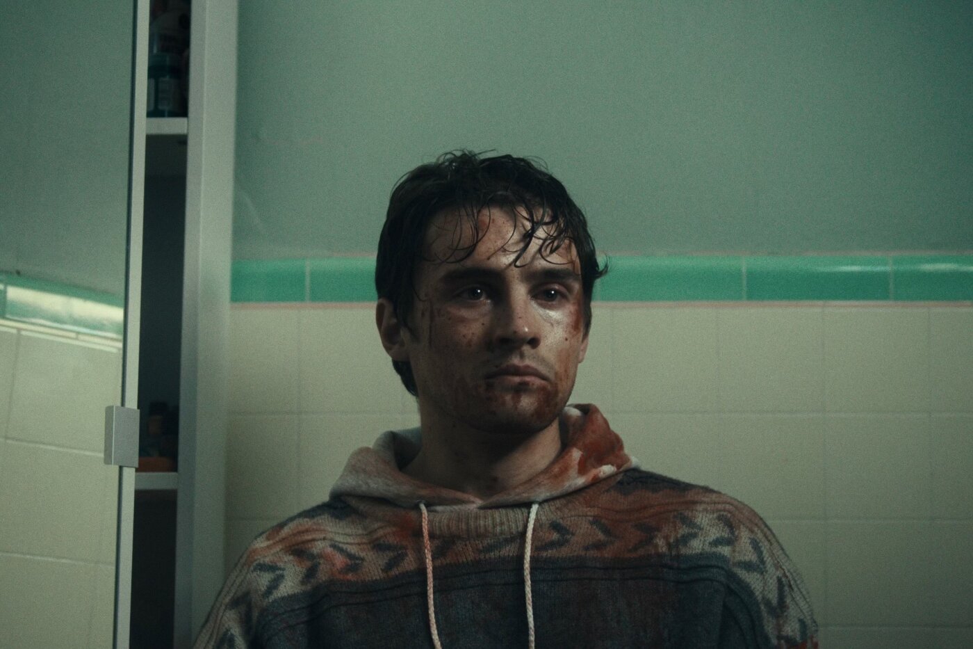 A man with dark, wet hair and dirt on his face stands in front of a tiled wall wearing a patterned zip-up hoodie.