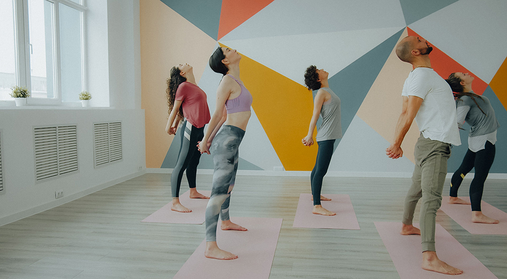 A group of people practicing a yoga or stretch pose in a studio with a colorful geometric mural on the back wall, all standing on mats and tilting their heads backward.