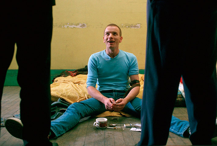 A high-angle shot from the movie Trainspotting showing Renton seated cross-legged on a mattress on the floor in a light-blue shirt and jeans with a tourniquet on his arm, framed between the dark silhouette legs of two figures standing over him.