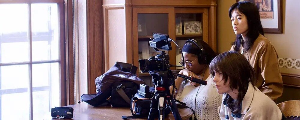 **Alt text:** A wide, cropped image showing a film crew gathered inside a sunlit room next to a large grid window, operating a camera set up on a tripod.