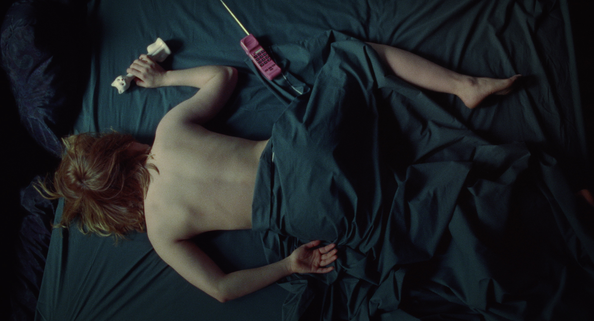 An overhead, high-angle shot showing a person lying face down on dark green bedsheets with their bare back exposed and dark sheets partially covering their legs.