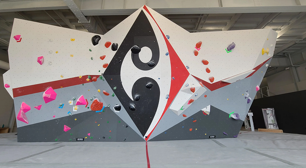 **Alt Text:** An indoor bouldering wall featuring a geometric design with grey, white, red, and black panels, a central black diamond logo, and colorful climbing holds scattered across the wall.