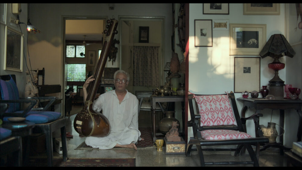 Still from the film Alaap showing a person sitting cross-legged on the floor playing a stringed instrument inside a room.