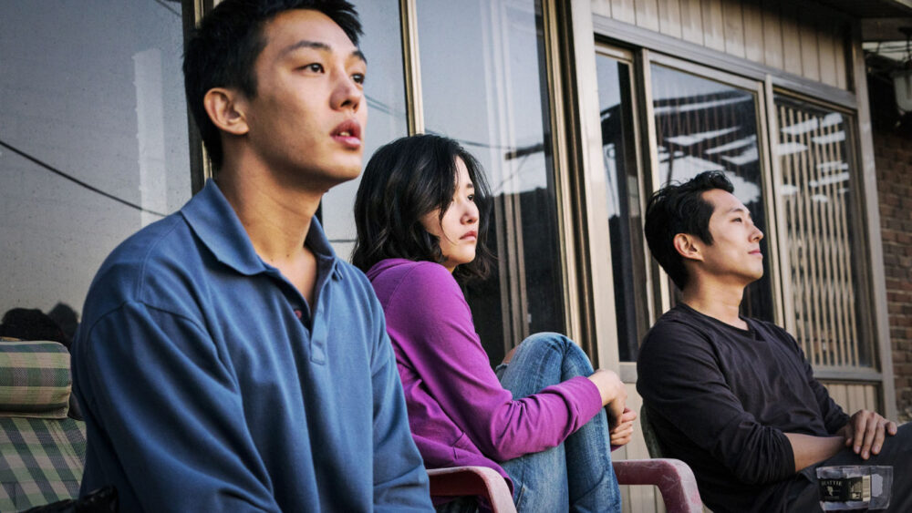 This image shows three actors from the 2018 South Korean psychological mystery drama film Burning, seated outdoors and looking off into the distance. From left to right: Steven Yeun (wearing a blue collared shirt), Jeon Jong-seo (in a purple top), and Yoo Ah-in (in a black long-sleeve shirt).