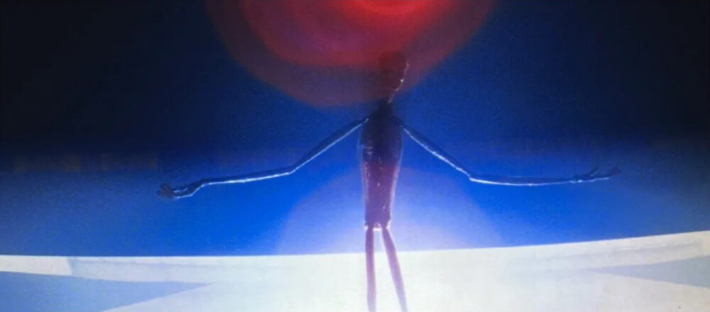 Still from the film Close Encounters of the Third Kind showing an alien figure with outstretched arms illuminated against a bright, glowing background.