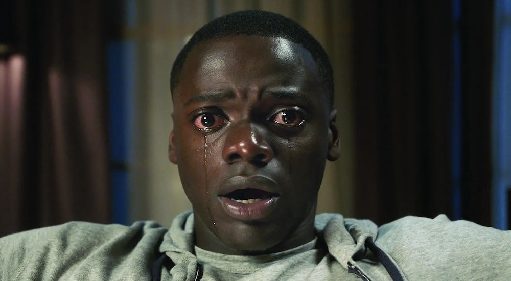 From the film Get Out, a medium close-up shot of Chris Washington (played by Daniel Kaluuya) sitting in a chair, experiencing the sunken place. He wears a light-colored sweatshirt and stares forward with wide, terrified eyes and tears welling up, his mouth slightly open in shock and distress.