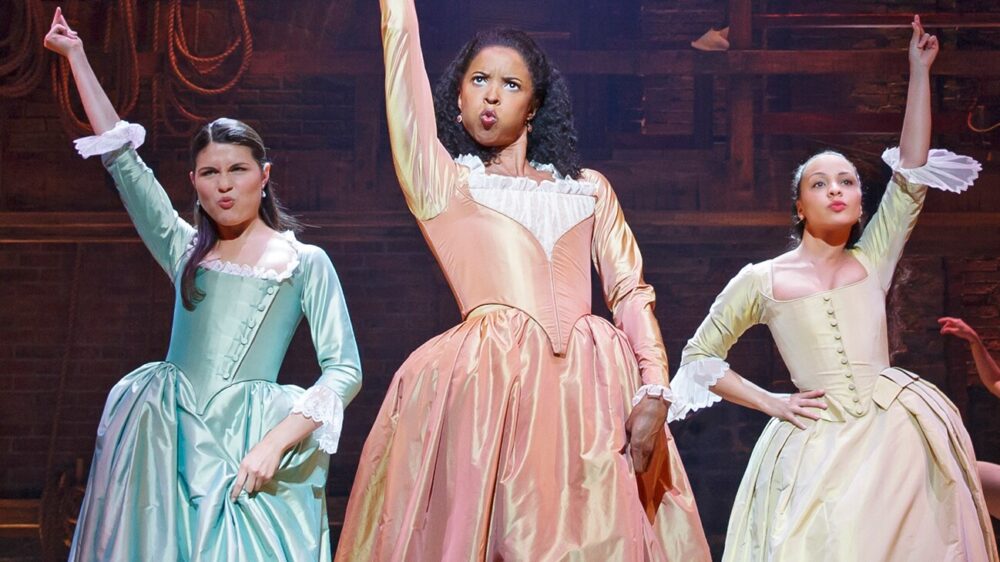 Production still from Hamilton showing the Schuyler sisters—Angelica in pink, and Eliza and Peggy in blue and green period dresses—raising their hands in performance.