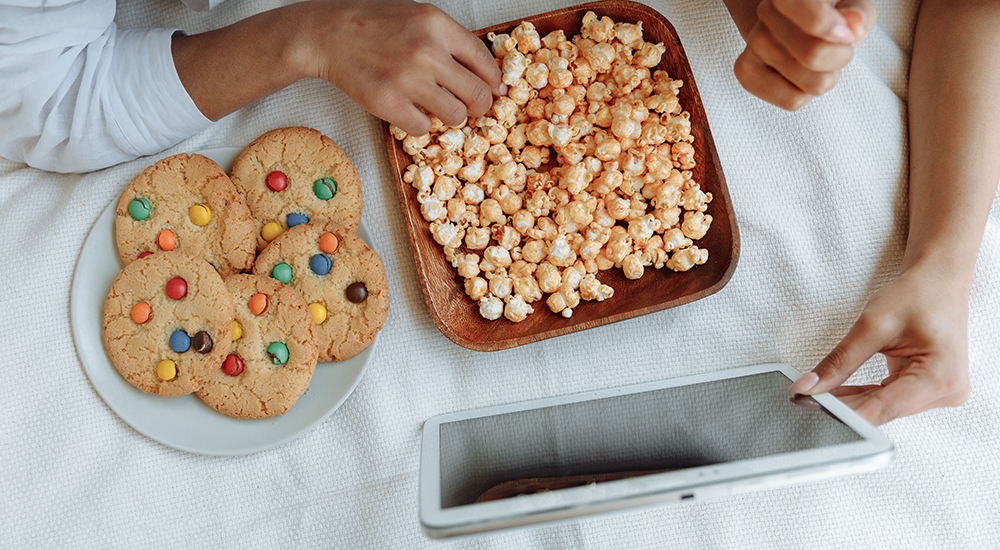 An overhead view of hands reaching for snacks around a digital tablet on a light surface, featuring a plate of chocolate button cookies and a wooden bowl of popcorn.