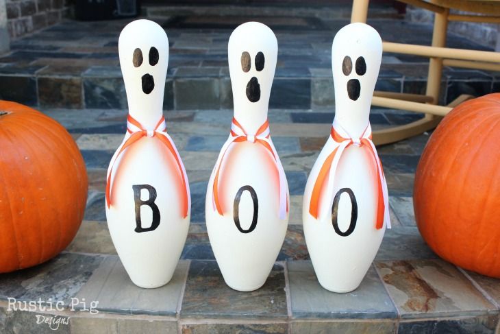 DIY Halloween craft featuring three painted bowling pins transformed into ghosts with faces, tied with orange ribbon, and spelling out the word "BOO" flanked by pumpkins.