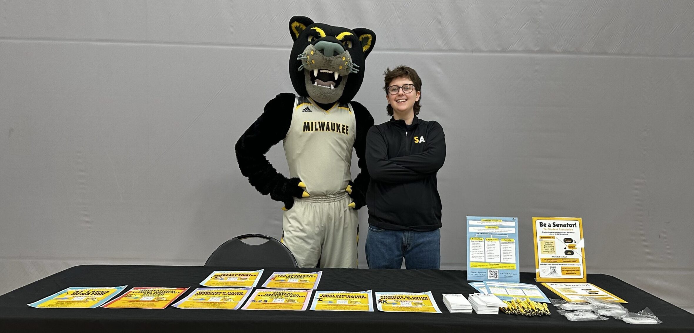 A promotional table is set up indoors with a black tablecloth displaying bright yellow and white flyers, brochures, pens, and small giveaway items. Behind the table stands the Milwaukee Panthers mascot, a black panther wearing a cream-colored basketball uniform with “MILWAUKEE” printed across the chest, alongside a person in a black long-sleeve shirt with “SA” printed on it. The backdrop is a plain gray wall, and the table signage includes phrases like “Be a Senator!” and information about student involvement opportunities, suggesting this is an event for student government or campus engagement at the University of Wisconsin-Milwaukee.