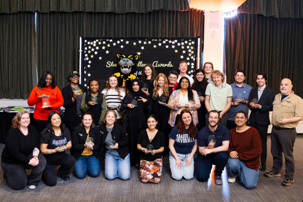 A group of approximately twenty people is standing and kneeling in rows inside a decorated event space. They are posing for a photo in front of a black backdrop adorned with gold and white star designs and text that reads “Student Stellar Awards.” The backdrop also features a mascot dressed in black with a gold accent, positioned near the center. Most individuals are holding small awards or trophies, indicating they are recipients. The setting has dark green curtains and bright lighting from the sides, creating a celebratory atmosphere. This image represents an award ceremony recognizing student achievements.