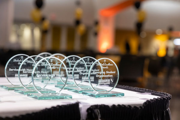 A close-up view of several clear glass awards arranged in a neat row on a white table with a black ruffled edge. Each award is circular and mounted on a rectangular glass base. The engraved text on the awards reads “Student Excellence Awards” with categories such as “Service with Heart” and “Student Emerging Organization,” along with the University of Wisconsin–Milwaukee logo. The background shows an event space with black and gold balloons and softly blurred tables and chairs, creating a festive atmosphere for the Student Excellence Awards ceremony.