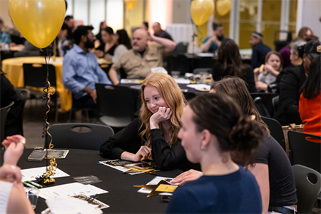 People are seated at round tables covered with black tablecloths in a large, well-lit event space with big windows in the background. Each table has gold and black decorations, including gold balloons tied to weights and printed programs or flyers. The attendees appear to be engaged in conversation or listening to something happening off-camera. The setting is part of the Student Stellar Awards, an event recognizing outstanding student achievements, with a celebratory atmosphere highlighted by the gold-themed decor.