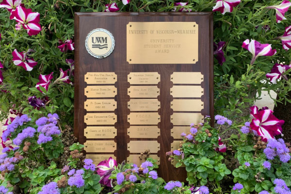 A wooden plaque with a polished finish is displayed outdoors among vibrant flowers, including purple and pink petunias and clusters of small lavender blooms. The plaque features a silver and black circular emblem at the top left with the letters “UWM” and a wave design, representing the University of Wisconsin–Milwaukee. Below the emblem, a large gold plate reads: “University of Wisconsin–Milwaukee, University Student Service Award.” Beneath this, multiple smaller gold plates are arranged in two columns, engraved with names and organizations recognized for the award. Some plates are blank, leaving space for future honorees. The plaque is surrounded by lush green foliage and colorful flowers, creating a celebratory and formal presentation.