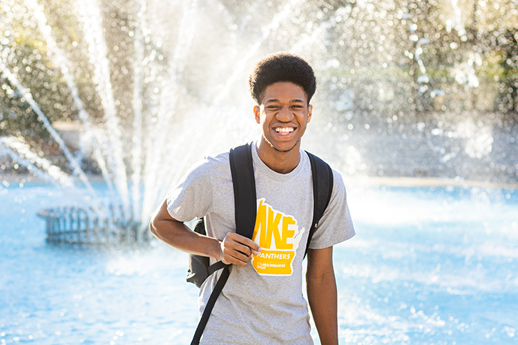 A smiling young black man wearing a UWM t-shirt standing in front of the UWM fountain