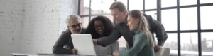 Four people gathered around a laptop at a table in a bright room with large industrial-style windows. One person is pointing at the laptop screen while others look on, suggesting collaboration or problem-solving.