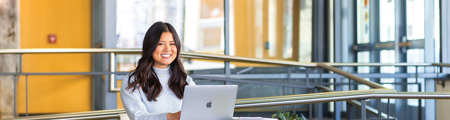 Student at a table in a gold room studying on their laptop and smiling at the camera