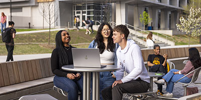 Three students sit outside, gathered around a laptop and laugh with each other.