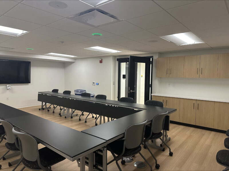 A modern conference or meeting room with a U-shaped arrangement of black tables and gray chairs on wheels. The room has light wood flooring, white walls, and a ceiling with recessed lighting panels. On the left wall, there is a large flat-screen monitor mounted above a small camera, suggesting video conferencing capability. The back wall features a set of wooden cabinets with a white countertop for storage. A black-framed glass door is open, leading to an adjacent space. The overall setting appears clean, professional, and designed for collaborative meetings or training sessions.