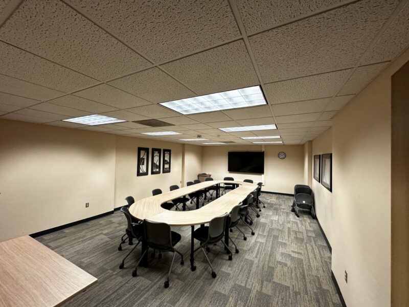 Medium-sized conference room with beige walls and a U-shaped arrangement of light wood tables in the center. Around 14 gray chairs with black bases are positioned along the tables, and two extra chairs are stacked against the right wall. The room has gray patterned carpet tiles and a ceiling with fluorescent light panels. On the far wall, a large flat-screen TV is mounted above a small cabinet, and a round wall clock hangs nearby. Three framed abstract artworks are displayed on the left wall, and another framed piece is on the right wall.