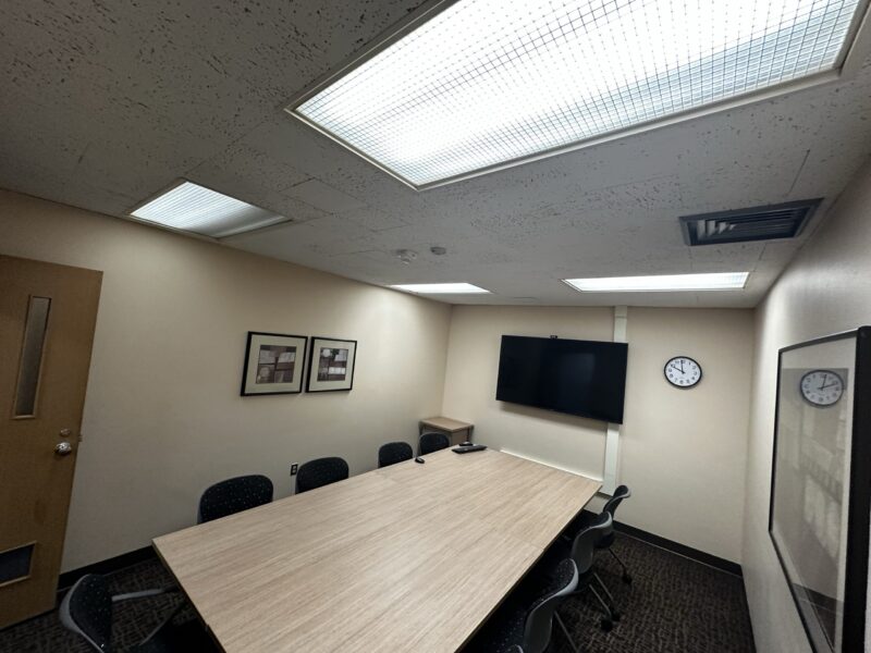 Small conference room with beige walls and a rectangular light wood table in the center, surrounded by eight black chairs with perforated backs. The room has a dark patterned carpet and a ceiling with fluorescent light panels. On the far wall, a large flat-screen TV is mounted above a small cabinet, and two round wall clocks are visible—one on the far wall and another on the right wall. Two framed abstract artworks hang on the left wall, and another framed piece is on the right wall near the door.