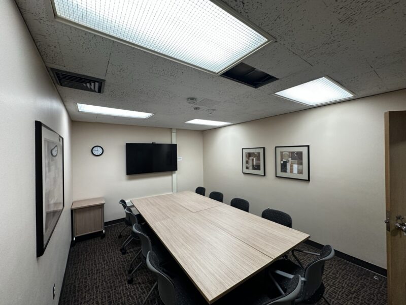 Small conference room with beige walls and a rectangular light wood table in the center, formed by two tables pushed together. Eight black chairs with perforated backs surround the table. The room has a dark patterned carpet and a ceiling with fluorescent light panels, one of which is missing its cover. On the far wall, there is a large flat-screen TV mounted above a small cabinet and a round wall clock. Two framed abstract artworks hang on the right wall, and another framed piece is on the left wall near the door.