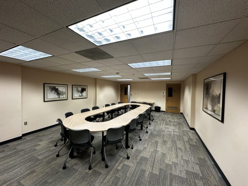 Large conference room with beige walls and a U-shaped arrangement of light wood tables in the center. About 14 gray chairs with black bases surround the tables. The floor is covered with gray patterned carpet tiles, and the ceiling has multiple fluorescent light panels. Two framed abstract artworks hang on the left wall, and another framed piece is on the right wall. At the far end of the room, there are two wooden doors, one with a small window panel. A trash bin is visible near the back wall.