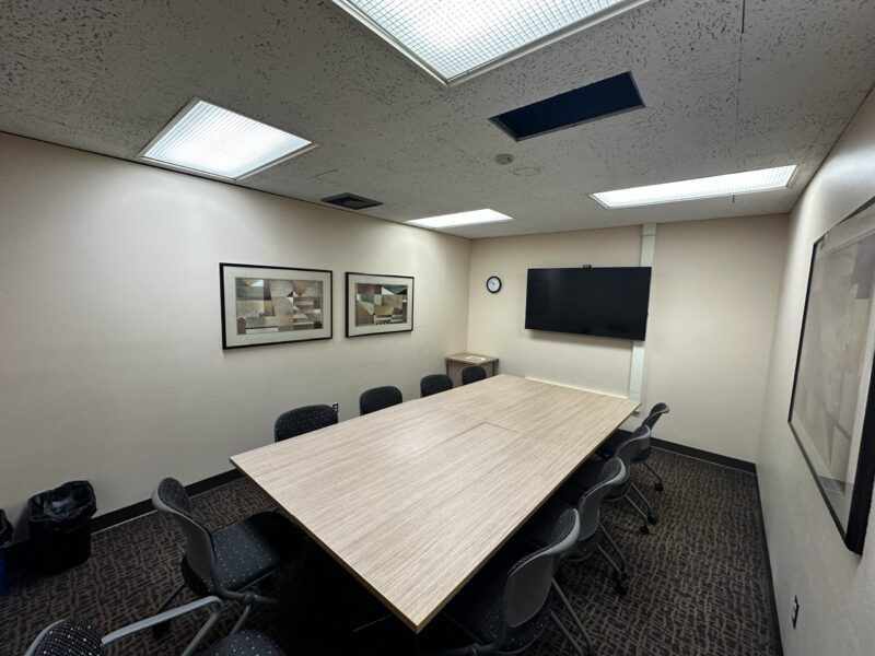 Medium-sized conference room with beige walls and a rectangular light wood table in the center, formed by two tables pushed together. Ten black chairs with perforated backs surround the table. The room has a dark patterned carpet and a ceiling with fluorescent light panels, one of which is missing its cover. On the far wall, there is a large flat-screen TV mounted above a small cabinet and a round wall clock. Two framed abstract artworks hang on the left wall, and another framed piece is on the right wall. A trash bin is visible in the left corner.