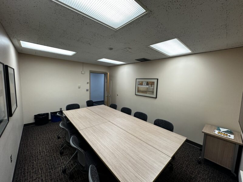 Small conference room with beige walls and a rectangular table made of light wood in the center. The table is formed by two smaller tables pushed together and surrounded by ten black chairs with perforated backs. The room has a patterned dark carpet and a ceiling with four fluorescent light panels. On the far wall, there is a framed abstract artwork and an open door leading to a hallway. A small rolling cart with a tray of items is positioned in the right corner, and two trash bins are visible in the left corner.