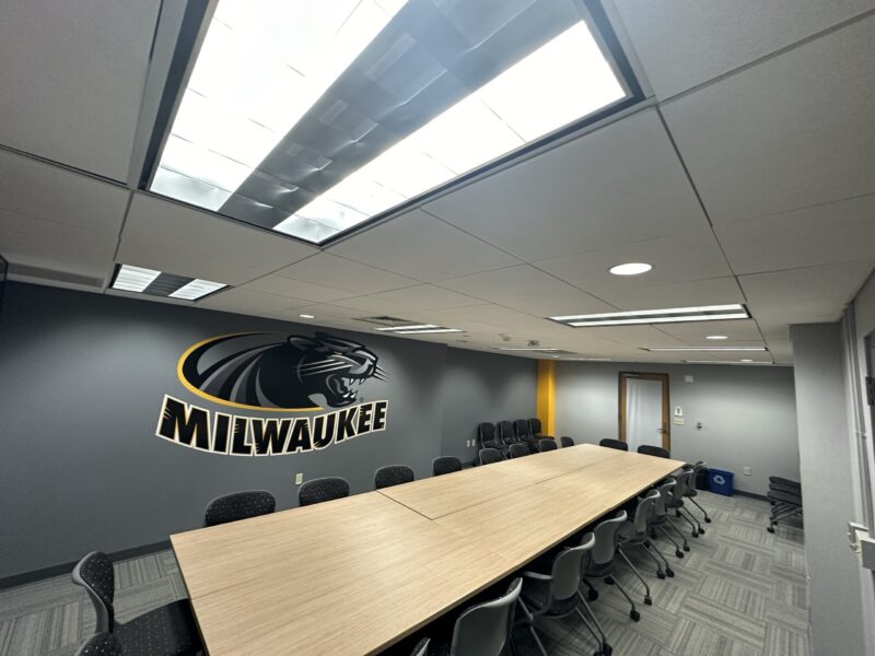 A modern conference room with gray walls and a patterned gray carpet. A long rectangular wooden table is centered in the room, surrounded by black chairs on wheels. The back wall features a large Milwaukee Panthers logo in black and gold. Bright fluorescent lights are recessed into the white ceiling. Additional chairs are stacked against the far wall near a wooden door, and a recycling bin is visible in the corner.