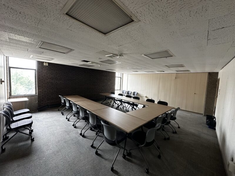 A dimly lit conference room with U-shaped tables made of light wood, surrounded by gray chairs on wheels. The room has gray carpet, a white textured ceiling with multiple recessed light panels, and beige paneled walls. Two tall windows on the left and far wall provide limited natural light and show greenery outside. Extra chairs are stacked along the left wall.
